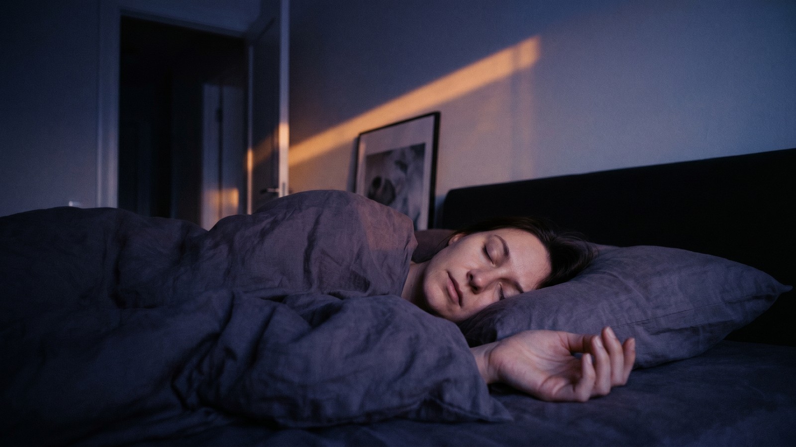 A person lying in bed in near-darkness, eyes softly closed, one hand resting open on the pillow, with a thin line of warm amber light crossing the wall