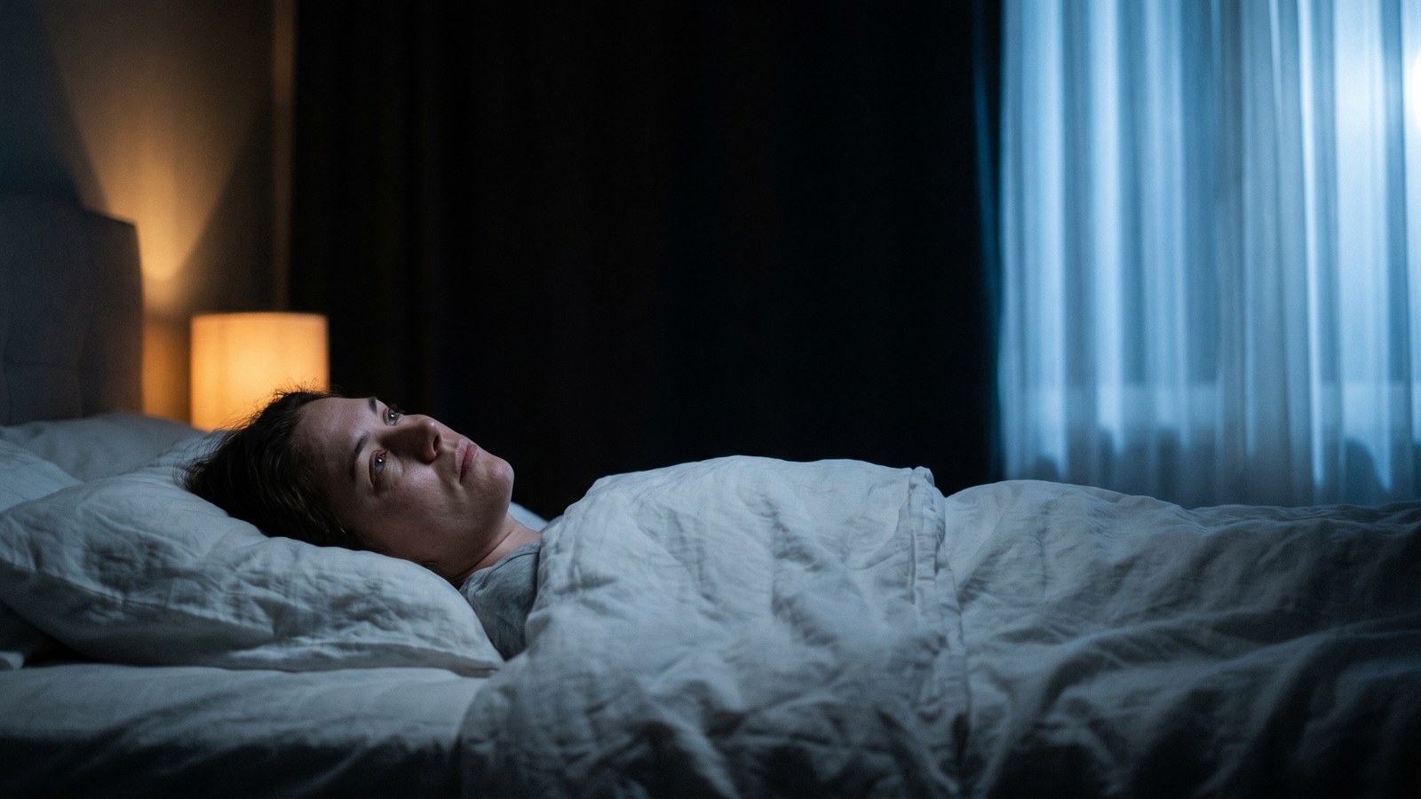 Person lying awake in a dimly lit bedroom, eyes open, staring at the ceiling with soft ambient light casting shadows across white bedsheets