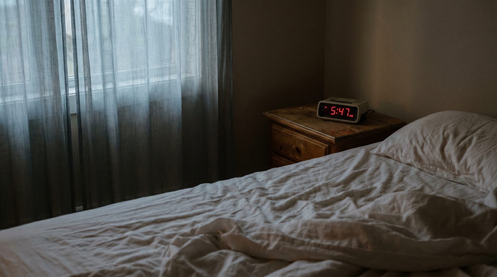 Rumpled bed sheets in dim early-morning light with a clock showing 5:47 AM on the bedside table
