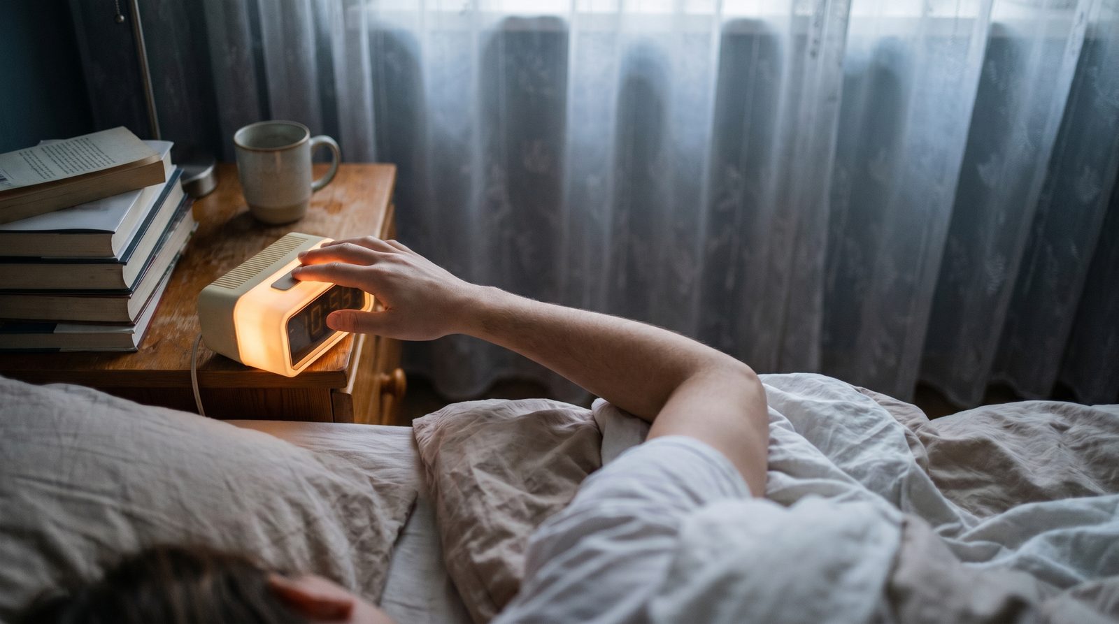 A hand reaching from tangled bedsheets to silence an alarm clock in soft grey-blue dawn light